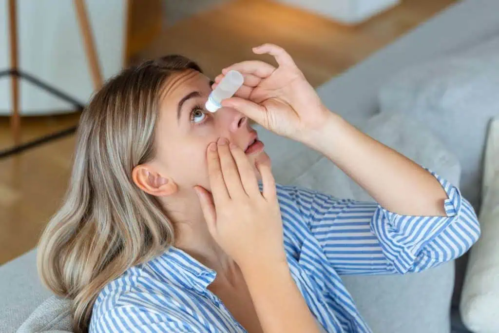 A person with blonde hair is seated on a couch using eye drops, following advice from an eye doctor in Suffolk County, NY. They tilt their head back, holding the bottle above their eye with one hand while gently pulling down their lower eyelid with the other. The person is wearing a striped shirt.