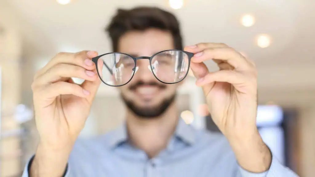 A man holds a pair of black-rimmed eyeglasses towards the camera, with a blurred background that hints at the quality eye care services in Suffolk County, NY. He is smiling, though his face isn’t clearly visible. The focus is on the glasses, showcasing their sleek design.
