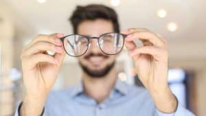 A man holds a pair of eyeglasses in front of him, smiling, with the glasses in focus and his face blurred in the background. The scene is brightly lit, highlighting professional eye care Suffolk County residents can trust.
