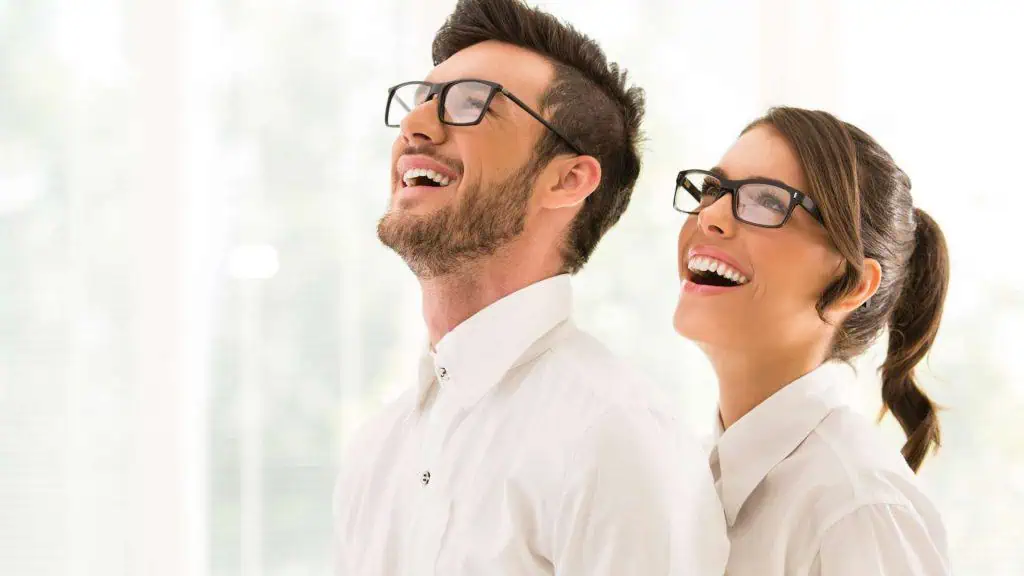 A man and a woman, both wearing glasses and white shirts, are standing close together, smiling and looking upwards, embodying the excellence of Suffolk County NY optometrists. The background is softly blurred, creating a bright and cheerful ambiance.