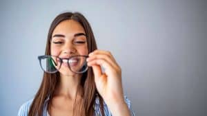 Smiling young woman with long brown hair holds a pair of eyeglasses in front of her face, looking at them—reflecting quality eye care Suffolk County residents trust. She stands against a plain light grey background.