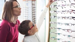 A woman and a young boy wearing glasses look at a display of eyeglass frames at an Optometrist Suffolk County, NY office. The boy points excitedly at a pair while the woman watches, both appearing interested in the selection.