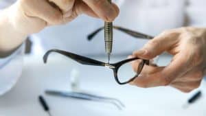 A person uses a small screwdriver to tighten or repair the hinge of eyeglasses, holding the glasses steady with one hand on a white surface—tools nearby suggest attention to detail and care, just like an Optometrist Suffolk County, NY provides.