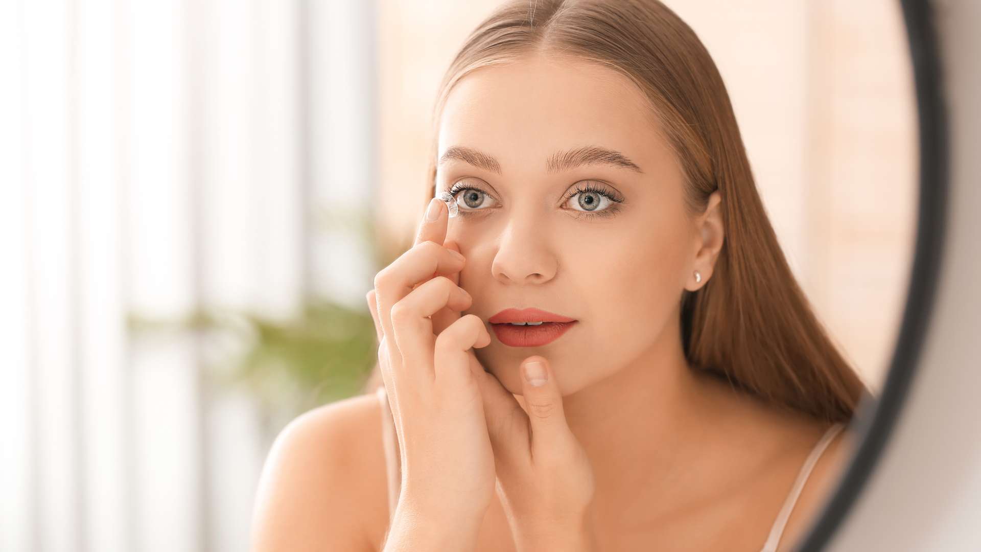 A young woman with long, straight hair is putting in a contact lens while looking into a mirror. She has light skin and blue eyes, showcasing the importance of eye care Suffolk County in a softly lit room while wearing a white top.