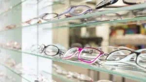 A display of various eyeglasses on glass shelves at a Suffolk eye optometrist. The glasses feature different frame styles and colors, including a distinctive pink pair in the center. The softly blurred background highlights the array of eyewear, showcasing eye care services in Suffolk County NY.