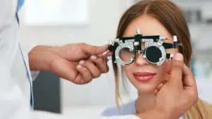 A woman with long hair sits in an eye exam chair, wearing a phoropter. An eye doctor from Suffolk County, NY, adjusts the device to test her vision in a clinical setting.