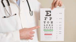 A Suffolk eye optometrist in a white coat, with a stethoscope around their neck, holds up an eye chart and points at a line. The chart displays letters of varying sizes, commonly used for eye exams and essential eye care services in Suffolk County, NY.