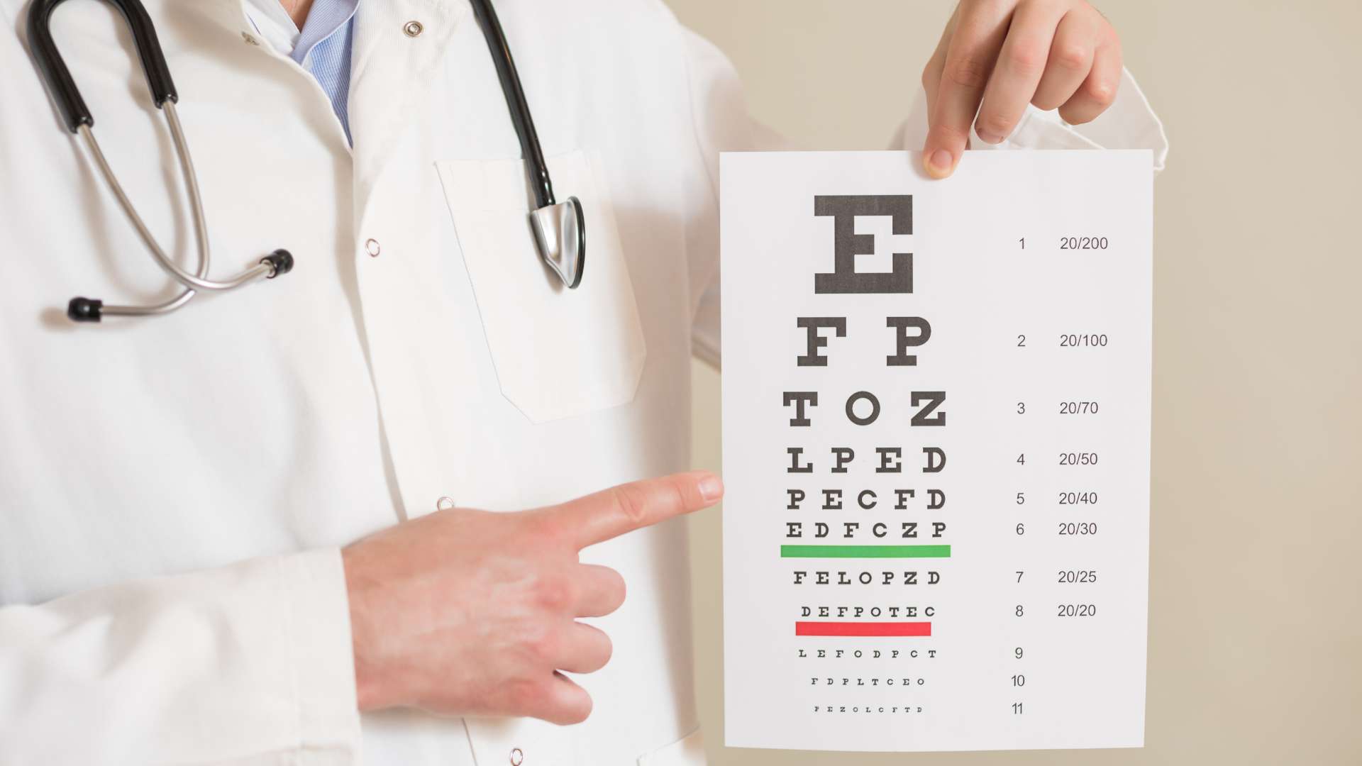 A doctor in a white coat holds and points to a standard eye chart with rows of letters for vision tests—ideal for anyone seeking expert eye care in Suffolk County. A stethoscope hangs around the Optometrist's neck.