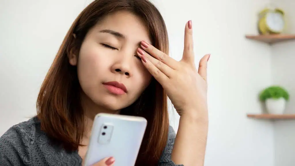 A woman with long hair closes her eyes, rubbing them wearily while holding a smartphone in the other hand. In the background, a small plant and a clock sit on a shelf. Perhaps she needs eye care services in Suffolk County, NY to ease her strain in this home or office setting.