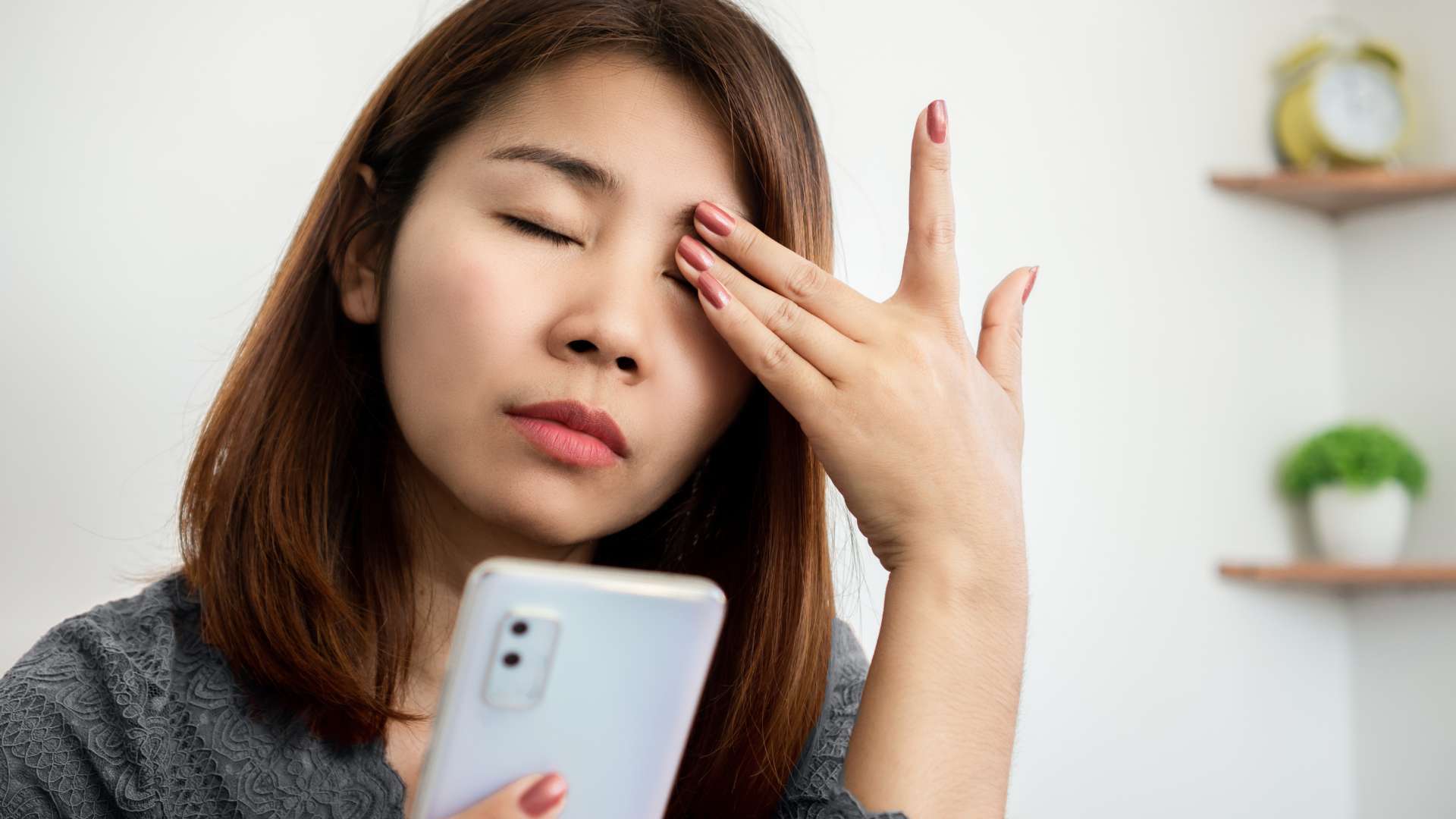 A woman in NY holds a smartphone and rubs her tired eye, eyes closed, in a bright indoor setting with blurred plants and a clock—reflecting the need for proper eye care Suffolk County residents can seek from an Optometrist Suffolk County.