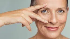 Close-up of a smiling person with light skin and blue eyes, gently touching their face with thumb and middle finger. The background is a neutral shade, reminiscent of a caring visit to an eye doctor in Suffolk County, NY.