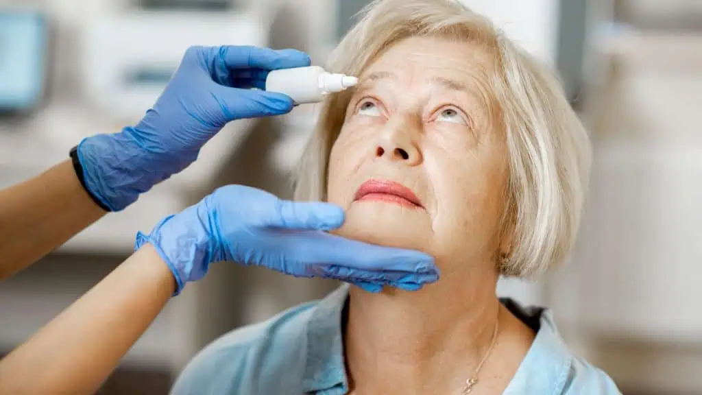 A woman with short blonde hair looks upward as an eye doctor administers eye drops. She appears calm, benefiting from the expert eye care services in Suffolk County, NY, while seated indoors.
