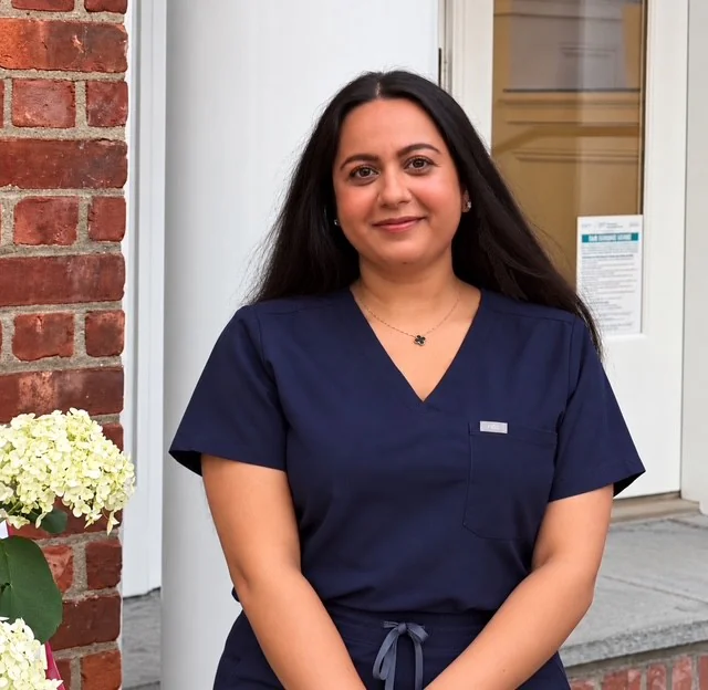 A smiling healthcare professional in navy blue scrubs stands outside a medical building.