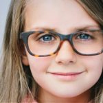 A close-up of a young child with long light brown hair, wearing large, dark-rimmed glasses, smiles gently at the camera—a charming portrait that highlights the importance of eye care in Suffolk County, NY. The background is a soft, blurred light gray.