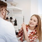 A young girl in a red plaid shirt smiles while sitting at an eye exam machine, with an Optometrist Suffolk County seated opposite her in a bright, modern clinic.