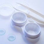 A pair of clear contact lenses, a white contact lens case with open lids, plastic tweezers, and two clear lids arranged on a white surface—ideal for those prioritizing eye care in Suffolk County, NY.