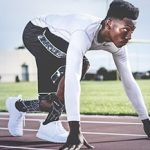 A male athlete wearing a white long-sleeve shirt, black shorts, and white running shoes is crouched at a starting line on an outdoor track, preparing to sprint. With a focused expression, he highlights the importance of eye care Suffolk County athletes trust.