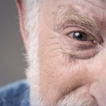 Close-up of an older man's face, focusing on his right eye and forehead, showing wrinkles and white facial hair. The background is blurred, and he appears to be smiling gently—reflecting trusted eye care Suffolk County, NY.