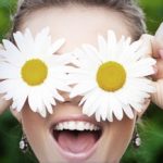 A smiling person holds two large daisies over their eyes, creating a playful and whimsical look. The green, blurred background adds charm—a fun reminder that quality eye care Suffolk County is always in sight.