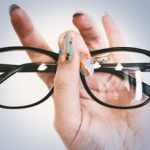 A hand with colorful, patterned nail art holds up black-rimmed eyeglasses against a light blue background, highlighting the importance of stylish vision and expert eye care in Suffolk County, NY.