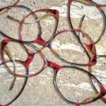 Several colorful eyeglass frames, perfect for eye care in Suffolk County, are arranged overlapping each other on a textured, sandy surface with white streaks running through the background.