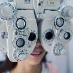 A person is sitting behind a phoropter, an eye exam device with multiple dials and lenses, as part of a vision test at an Optometrist Suffolk County office. The device obscures most of their face.