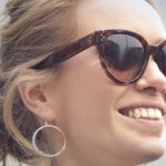A smiling woman with light brown hair in a loose bun, wearing large tortoiseshell sunglasses and a large hoop earring, highlights the stylish side of eye care in Suffolk County, NY.