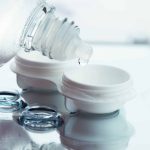 A close-up of a contact lens case with the lid open, two contact lenses beside it, and a bottle dispensing solution above—all reflecting below. Perfect illustration for those seeking eye care Suffolk County, NY.