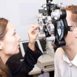 An optometrist in Suffolk County adjusts a phoropter in front of a man's face during an eye exam in a clinical NY setting. The man is seated while the optometrist is focused on providing expert eye care Suffolk County residents trust.